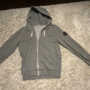Grå Stone Island hoodie med dragkedja - Snygg grå hoodie från Stone Island med klassisk logga på ärmen. Tröjan har huva med snörning, hel dragkedja framtill och ribbade muddar. Tillverkad i mjuk bomull för en skön och avslappnad passform. Perfekt för dig som gillar streetwear och stilren design. Muddarna har varit sönder men de är ihop sydda som ni ser på bilden , muddarna är väldigt lätt bytna. Sedan är det något mer små fel som ni ser på bilderna.