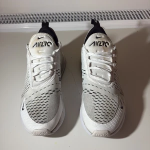 Nike Air Max 270 vita sneakers - Nike Air Max 270 sneakers i vit med svarta detaljer och stor synlig luftbubbla i hälen. Ovandelen är i ventilerande syntetmaterial med snörning och svart logga på sidan. Snygg, sportig design med rund tå och platt sula. Perfekt för dig som gillar streetwear och komfort.