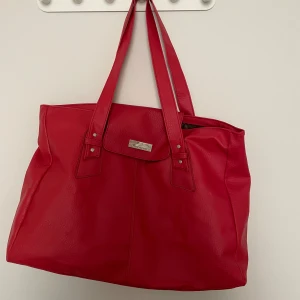 Röd axelväska i skinnimitation - Stor och rymlig röd axelväska med långa handtag och silverfärgad detalj framtill. Väskan har en stilren design och är tillverkad i fejkskinn