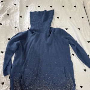 Blå polotröja med glitterdetaljer - Mysig blå polotröja med hög, ribbad krage och långa ärmar. Tröjan har två fickor framtill och är dekorerad med glittriga stenar nertill för en extra cool touch. Perfekt för dig som vill sticka ut lite extra under kyliga dagar.