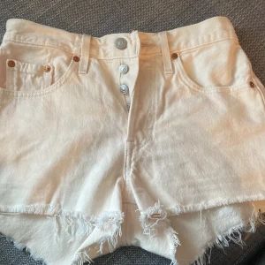 Vita Levi's 501 jeansshorts med rå kant - Säljer ett par vita Levi's 501 jeansshorts med fransig kant. Klassisk femficksmodell med knappar framtill och Levi's läderpatch bak. Perfekta för sommaren och riktigt snygga till sneakers eller sandaler. Skulle säga att de är ljus beige/rosa 💗