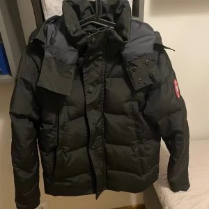 Svart pufferjacka från Canada Goose - Svart pufferjacka från Canada Goose med huva och klassisk logga på insidan. Jackan har dragkedja och tryckknappar framtill, samt flera praktiska fickor. Perfekt för kalla vinterdagar och har en snygg, stilren look.