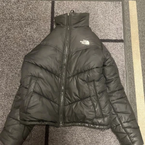 Svart pufferjacka från The North Face - Svart pufferjacka från The North Face med hög krage och synlig dragkedja framtill. Jackan har ett quiltat mönster och två sidofickor. Perfekt för kalla dagar med sin dunfyllda insida och stilrena look. Loggan är broderad i vitt på bröstet.