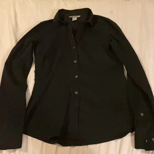 Svart skjorta från H&M, stl 34 - Klassisk svart skjorta från H&M i storlek 34. Skjortan har lång ärm, knappar framtill och vid manschetten. Materialet är en mix av bomull, polyester och lite elastan för extra komfort. Perfekt till en clean och stilren look.Figur sydd.