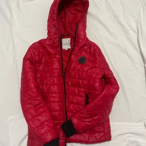 Röd Moncler pufferjacka  - Säljer en röd pufferjacka från Moncler med huva och svarta detaljer. Jackan har quiltad design, dragkedja framtill och ett Moncler-märke på bröstet. Insidan har en cool serietidningsetikett och jackan är tillverkad i polyester. Perfekt för dig som vill ha en snygg och varm jacka.