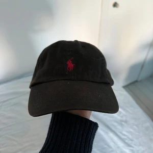 Svart keps från Polo Ralph Lauren - Svart keps från Polo Ralph Lauren med klassisk böjd skärm och röd broderad logga framtill. Tillverkad i bomull och har ventilerande hål upptill. Perfekt accessoar för en avslappnad och sportig look.