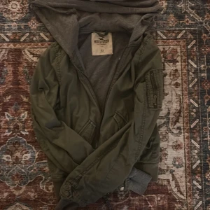 Grön bomberjacka från Hollister XS - Snygg grön bomberjacka från Hollister i storlek XS. Jackan har en mjuk grå huva, ribbade muddar och dragkedja framtill. Fickor med knappstängning och en cool ficka med dragkedja på ärmen. Perfekt för dig som gillar streetstyle och vill ha en chill look.