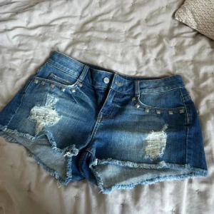 Blå jeansshorts med nitar och slitningar - Snygga blå jeansshorts med råa kanter, slitna detaljer och coola silverfärgade nitar vid fickorna. Klassisk femficksmodell i denim med fransig nederkant och slitningar både fram och bak. Perfekta för en chill sommarlook.