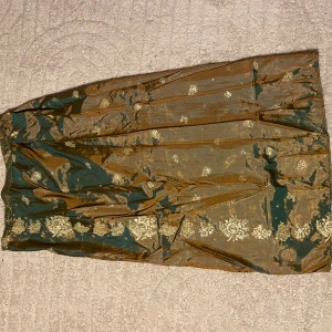 Grön och guld långkjol i siden - Unik långkjol i grönt och guldigt siden med vackra blommiga mönster. Kjolen har en glansig finish och ett elegant fall, perfekt för dig som vill sticka ut med en klassisk och lyxig look. Resår i midjan för skön passform. Egensydd.