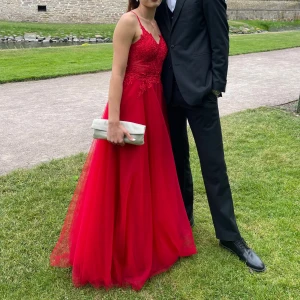 Röd balklänning med spetsdetaljer - Säljer min röda balklänning med tunna axelband och v-ringad spetsliv. Kjolen är i flera lager av tyll som ger en riktigt drömmig känsla, med en slits framtill på vänster sida . Perfekt för bal eller student! Klänningen har en figurnära överdel och en voluminös kjol som ger en elegant siluett. Endast använd en gång!