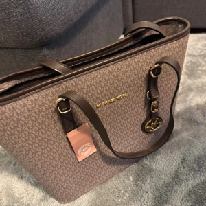 Brun axelväska från Michael Kors - Snygg brun axelväska från Michael Kors med MK-mönster över hela väskan. Väskan har guldfärgade detaljer, justerbara remmar och flera fack med dragkedja. Perfekt storlek för att få plats med allt du behöver och har en lyxig känsla med logotypen framtill.