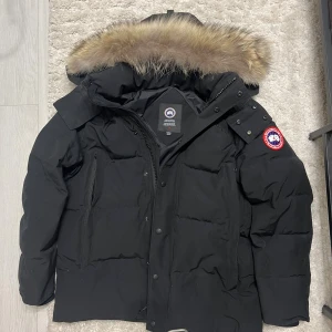 Svart wyndham Canada Goose M - Svart dunjacka från Canada Goose med ikonisk röd och vit patch på ärmen. Jackan har en stor huva med fluffig päls, dragkedja och knappar framtill samt praktiska fickor. Perfekt för kalla vinterdagar och har en clean, klassisk look.