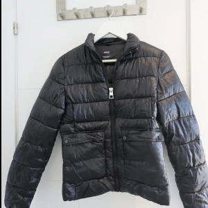 Svart pufferjacka från MANGO, S - Svart pufferjacka från MNG i storlek S. Jackan har quiltad design, två fickor med knappstängning framtill och dragkedja. Materialet är glansigt och syntetiskt, perfekt för kyliga dagar. Ståkrage och långärmad modell ger extra värme och stil.