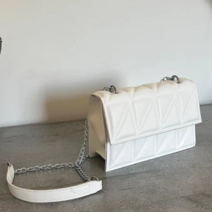 Vit quiltad axelväska med kedja - Snygg vit axelväska i quiltad design med geometriskt mönster. Väskan har en silverfärgad kedjerem och är tillverkad i skinnimitation. Perfekt storlek för det viktigaste och ger en clean look till din outfit.
