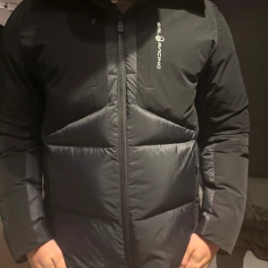 Svart Sail Racing pufferjacka med huva - Säljer en svart pufferjacka från Sail Racing med huva och snyggt logotryck på bröstet. Jackan har dragkedja framtill, quiltad design och lång ärm. Materialet är polyester och syntet, perfekt för kalla dagar. Passformen är normal och jackan har en modern look.