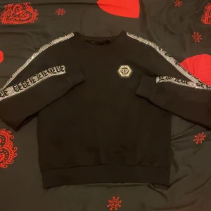 Svart Philipp Plein sweatshirt med strass - Svart sweatshirt från Philipp Plein med glittrande strassdetaljer längs ärmarna och ett stort PP-emblem på bröstet. Tröjan har rund hals och långa ärmar, perfekt för dig som gillar statement-plagg och exklusiv streetstyle.