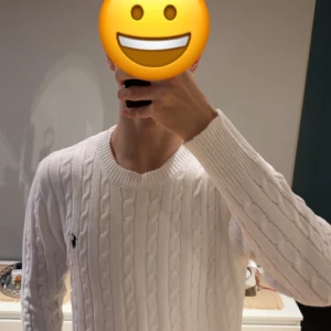 Vit kabelstickad tröja från Polo Ralph Lauren - Snygg vit kabelstickad tröja från Polo Ralph Lauren med klassisk rund hals och svart broderad logga på bröstet. Tröjan har långärmad passform och ribbade muddar vid ärmslut och hals. Perfekt för dig som gillar stilrena och tidlösa plagg.