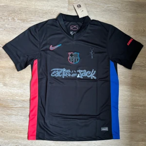 Svart FC Barcelona x Nike fotbollströja - Unik svart FC Barcelona fotbollströja från Nike med coola grafiska detaljer och Cactus Jack-tryck på bröstet. Tröjan har korta ärmar, röd och blå panel på sidorna, samt ett broderat FCB-märke med neonröd och blå kant. Tillverkad i lätt och ventilerande polyester.