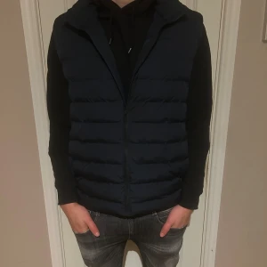 Mörkblå dunväst från Selected Homme - Snygg mörkblå dunväst från Selected Homme med quiltad design och dragkedja framtill. Västen har en stilren look och passar perfekt över en hoodie eller tröja. Materialet är polyester och västen har en normal passform.