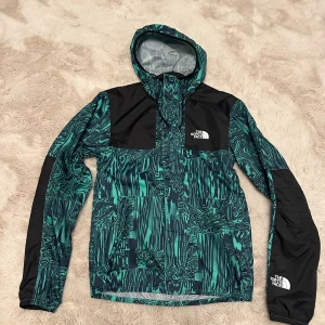 The North Face regnjacka XS - Snygg vindjacka från The North Face i storlek XS. Jackan har ett unikt grönt och svart mönster, huva med snörning och logga på bröstet och ärmen. Tillverkad i lätt polyester, perfekt för blåsiga dagar. Stängs med dragkedja och tryckknappar framtill.