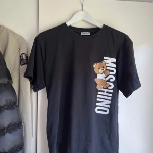 Svart Moschino t-shirt med björntryck - Cool svart t-shirt från Moschino med stort vertikalt Moschino-tryck och en björn på framsidan. Klassisk rund hals och korta ärmar. Tillverkad i mjuk bomull som är skön mot huden. Perfekt för dig som gillar streetstyle och statement-plagg.