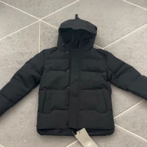 Svart pufferjacka från Canada Goose - Svart pufferjacka från Canada Goose med huva och stora fickor framtill. Jackan har en stilren look med raka linjer och diskreta detaljer. Perfekt för kalla dagar med sin tjocka vaddering och klassiska design. Märket syns både på insidan och på taggarna.