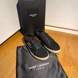 Svarta Saint Laurent sneakers - Snygga svarta sneakers från Saint Laurent med vit logga på sidan och kontrasterande beige sula. Skorna har klassisk rund tå, snörning och är tillverkade i en mix av skinn och textil. Perfekta för dig som gillar stilrena och exklusiva sneakers. Pris går att diskutera.