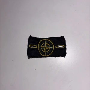 Stone Island patch svart/gul - Säljer en klassisk svart patch från Stone Island med det ikoniska kompassloggan broderad i gult och grönt. Märket har två knapphål på sidorna och passar perfekt att fästa på jacka eller tröja för en streetwear-look.