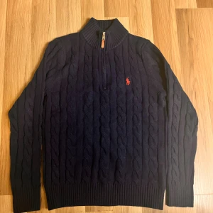 Mörkblå stickad tröja Polo Ralph Lauren - Snygg mörkblå stickad tröja från Polo Ralph Lauren med klassiskt kabelmönster och röd broderad logga på bröstet. Tröjan har half zip, ribbad krage och långa ärmar. Perfekt för dig som gillar stilren och tidlös design.