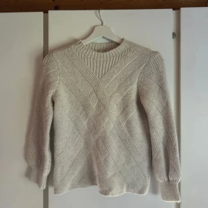 Beige stickad tröja med ribbad struktur - Säljer en beige stickad tröja med snyggt ribbad mönster i korsande linjer. Tröjan har rund hals och långa ärmar med lätt puff vid mudden. Perfekt för dig som gillar en enkel men ändå unik look.