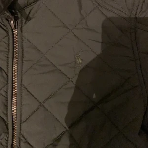 Olivgrön quiltad jacka Polo Ralph Lauren - Snygg olivgrön quiltad jacka från Polo Ralph Lauren med dragkedja och två stora fickor framtill. Jackan har ståkrage med spänne, långärmad design och diskret broderad logga på bröstet. Perfekt för höst och vinter när du vill ha en clean och klassisk look.