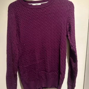 Lila kabelstickad tröja - Säljer en lila kabelstickad tröja med rund halsringning och långa ärmar. Tröjan har en klassisk, ribbad kant vid hals, ärmslut och nederkant. I märket Hampton republic 