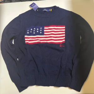 Mörkblå stickad tröja Polo Ralph Lauren - Mörkblå stickad tröja från Polo Ralph Lauren med amerikansk flagga i rött, vitt och blått på bröstet. RL-broderi i rött nedanför flaggan. Klassisk rund halsringning och ribbade muddar. Perfekt för dig som gillar preppy stil.priset kan diskuteras vid snabb affär.