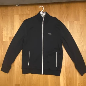 Mörk blå Hugo Boss kofta - Mörkblå kofta från BOSS med vit dragkedja och vita detaljer vid fickorna. Koftan har ståkrage med snygga vita och svarta ränder på insidan. Klassisk och stilren design med BOSS-logga på bröstet. Perfekt för dig som gillar sportig och clean look.