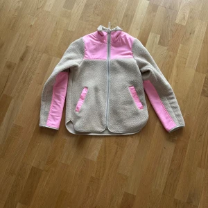 Beige teddyjacka med rosa detaljer Röhnisch - Säljer en beige teddyjacka från Röhnisch i storlek XS med ljusrosa paneler på axlar, ärmar och fickor. Jackan har hel dragkedja framtill, hög krage och mjukt fleece-material som är perfekt för kyliga dagar. Snygg och sportig look med kontrastfärger.