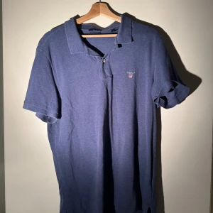 Blå pikétröja från GANT - Snygg blå pikétröja från GANT i storlek XL. Klassisk krage, korta ärmar och två knappar framtill. Tröjan har en liten broderad GANT-logga på bröstet och är gjord i mjuk bomull. Perfekt för en chill och stilren look.