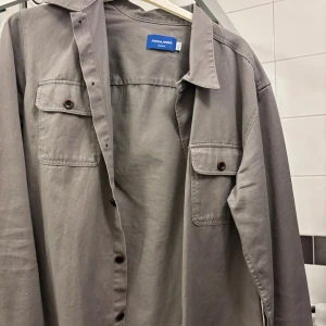 Grå overshirt från Jack & Jones - Snygg grå overshirt från Jack & Jones Originals med klassisk krage och två bröstfickor med knappar. Skjortan har långa ärmar och är tillverkad i mjuk bomull. Perfekt att bära som lager över en t-shirt för en chill look.