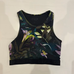 Blommig sporttopp från OTP - Snygg sporttopp från OTP med blommigt mönster i grönt, lila, gult och svart. bred resår nertill och har en hög halsringning. Perfekt för träning och ger en cool vibe med sitt färgstarka tryck.