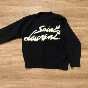 Svart Saint Laurent stickad tröja L - Svart stickad tröja från Saint Laurent i storlek L. Tröjan har ett stort vitt Saint Laurent-tryck framtill i en cool, suddig stil. Klassisk rund halsringning och ribbade muddar vid ärmslut och nederkant. Perfekt för dig som gillar exklusiva och trendiga plagg.