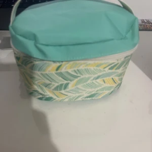 Turkos handväska med bladmönster - Snygg liten handväska i turkos med bladmönster i grönt och gult. Väskan har ett mjukt tygmaterial och praktisk dragkedja runt locket. Perfekt för att bära småsaker och har ett fräscht, somrigt utseende.