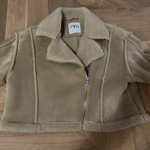 Beige teddyjacka från Zara - Säljer en beige teddyjacka från Zara med mjuk fuskpäls på krage, ärmslut och nederkant. Jackan har en croppad passform och cool asymmetrisk dragkedja framtill. Materialet är mockaliknande syntet med fluffiga detaljer som ger en riktigt mysig vibe.