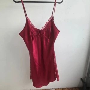 Röd satin klänning med spetsdetaljer - Snygg röd kort natt klänning i satin med tunna axelband som går att justera och v-ringning. Klänningen har spetsdetaljer vid bysten och längs sidorna samt en liten rosett framtill. 