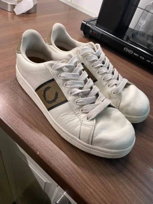 Fred Perry vita sneakers strl 41 - ANVÄNDA EN GÅNG. Snygga vita sneakers från Fred Perry med beige och svart detalj på sidan och klassisk lagerkrans-logga. Skorna har rund tå, platt sula och snörning. Ovandelen är i skinn och insidan är mjukt fodrad för extra komfort. Perfekt för dig som gillar stilrena och sportiga sneakers.