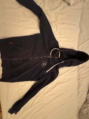 Mörkblå hoodie från Race Marine - Snygg mörkblå hoodie från Race Marine med dragkedja och huva. Tröjan har vita och svarta snören, två fickor fram och ett litet tryck på bröstet. Perfekt för chill dagar och sportiga outfits.