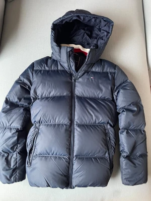 Mörkblå pufferjacka Tommy Hilfiger - Snygg mörkblå pufferjacka från Tommy Hilfiger med huva och dragkedja framtill. Jackan har två sidofickor med dragkedja och klassisk quiltad design. Insidan har en cool röd och vit rand längs kragen. Perfekt för kalla dagar och riktigt street.