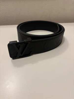 Svart Louis Vuitton skärp i skinn - Säljer ett svart Louis Vuitton bälte i skinn med präglade LV-loggor och monogram längs hela remmen. Bältet har en stilren, matt svart LV-spänne som ger en lyxig vibe. Perfekt för dig som vill ha en clean och exklusiv accessoar.