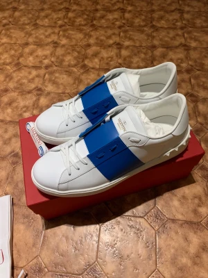 Valentino Garavani Open sneakers vit/blå - Snygga Valentino Garavani Open sneakers i vitt skinn med en bred blå läderpanel över snörningen. Rund tå, platt sula med mönster och klassisk logga på plösen. Kommer med originalförpackning och dustbag. Perfekt för dig som gillar exklusiva och stilrena sneakers.