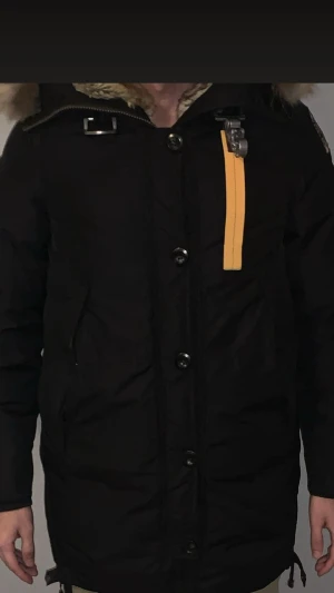 Svart Dam parkasjacka med päls från Parajumpers - Svart parkasjacka från Parajumpers med gul detalj vid halsen och stor huva med fluffig päls. Jackan har knappar och dragkedja framtill, samt praktiska fickor på sidorna. Perfekt för kalla dagar med sin långa passform och varma material. 