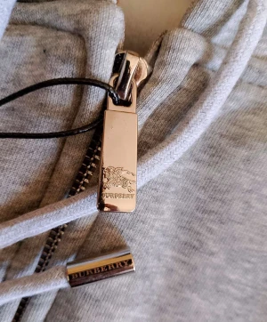 Grå hoodie från Burberry med dragkedja - Snygg grå hoodie från Burberry med klassisk rutig insida i huvan och broderad ryttare på bröstet. Tröjan har dragkedja framtill, snörning i huvan och ribbade muddar. Metall-detaljer med Burberry-logga på snörändarna ger en lyxig touch.