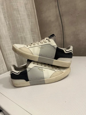 Svarta och vita sneakers från Pepe Jeans - Snygga sneakers från Pepe Jeans i svart och vitt med grå detaljer. Skorna har rund tå, platt sula och snörning framtill. Materialet är en mix av skinn och syntet, med en stilren blockdesign på sidorna och Pepe Jeans-logga i guld. Perfekt för dig som gillar clean och modern stil.